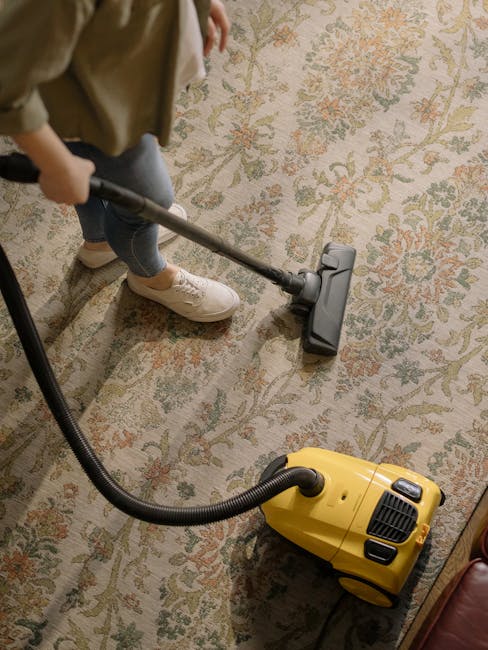 A woman with brown hair tied back, dressed in a light striped shirt and jeans, is kneeling on a patterned rug in a living room, engaged in surface cleaning with a blue cloth. The room features a dark 