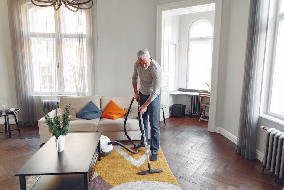 A person kneeling on a decorative patterned area rug in a residential setting, operating a yellow and black portable vacuum cleaner with a flexible hose. The individual is wearing a beige jacket and blue jeans, focusing on cleaning the carpet surface. The room features warm lighting, and the carpet appears clean, indicating effective surface cleaning and maintenance, consistent with professional domestic cleaning services offered by Carpet Cleaning W8 for period homes in Holland Park Avenue, W8.