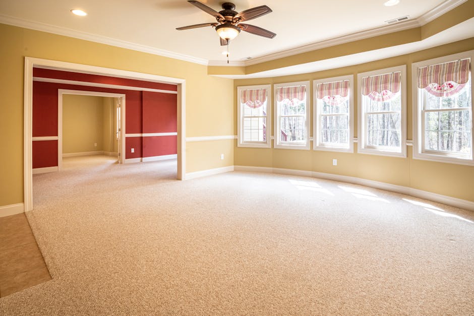 A bright, spacious living room featuring a soft beige carpet that appears clean and well-maintained, with walls painted in a light yellow shade and decorative white trim. Multiple large windows with p