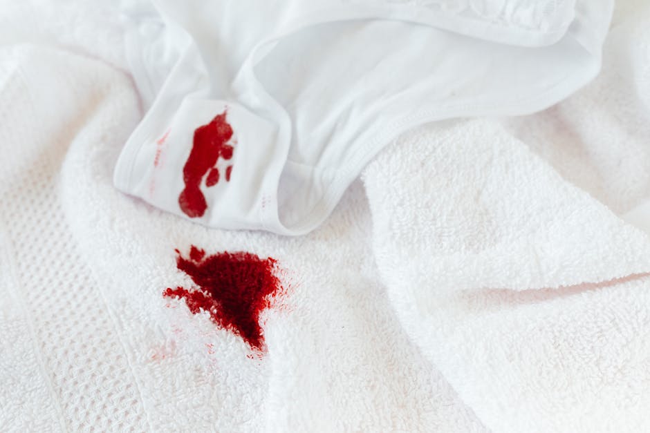 A close-up image of a wine glass lying on its side with red wine spilled onto a white surface, creating a colorful stain. The glass is made of clear glass with a slender stem, and the deep red wine in