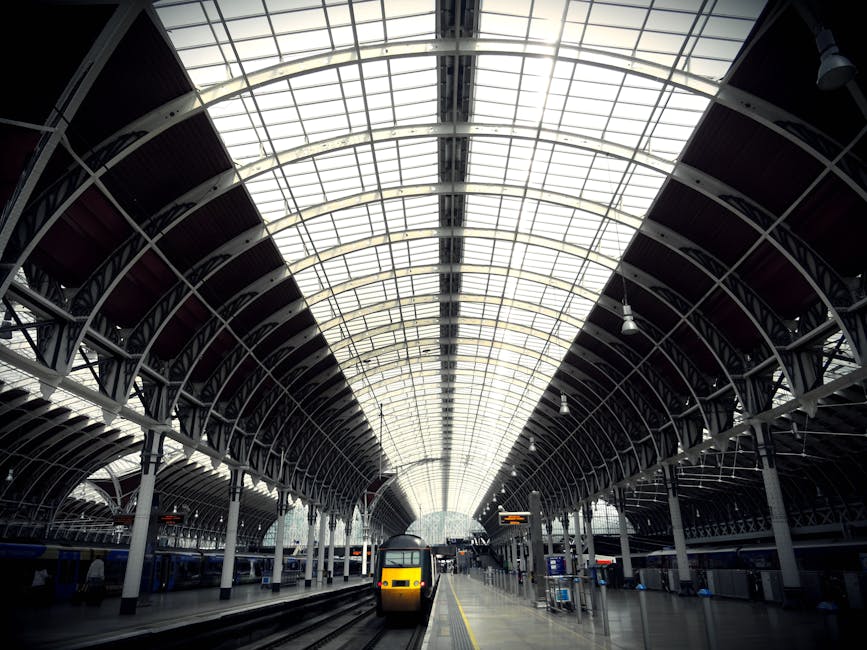 Interior view of a modern train station with a high, arched glass ceiling supported by metal framework, allowing natural light to illuminate the platform below. The platform features tiled flooring, c