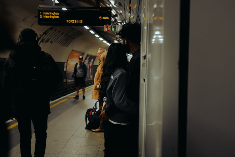 Interior view of a modern train station with a high, arched glass ceiling supported by metal framework, allowing natural light to illuminate the platform below. The platform features tiled flooring, c